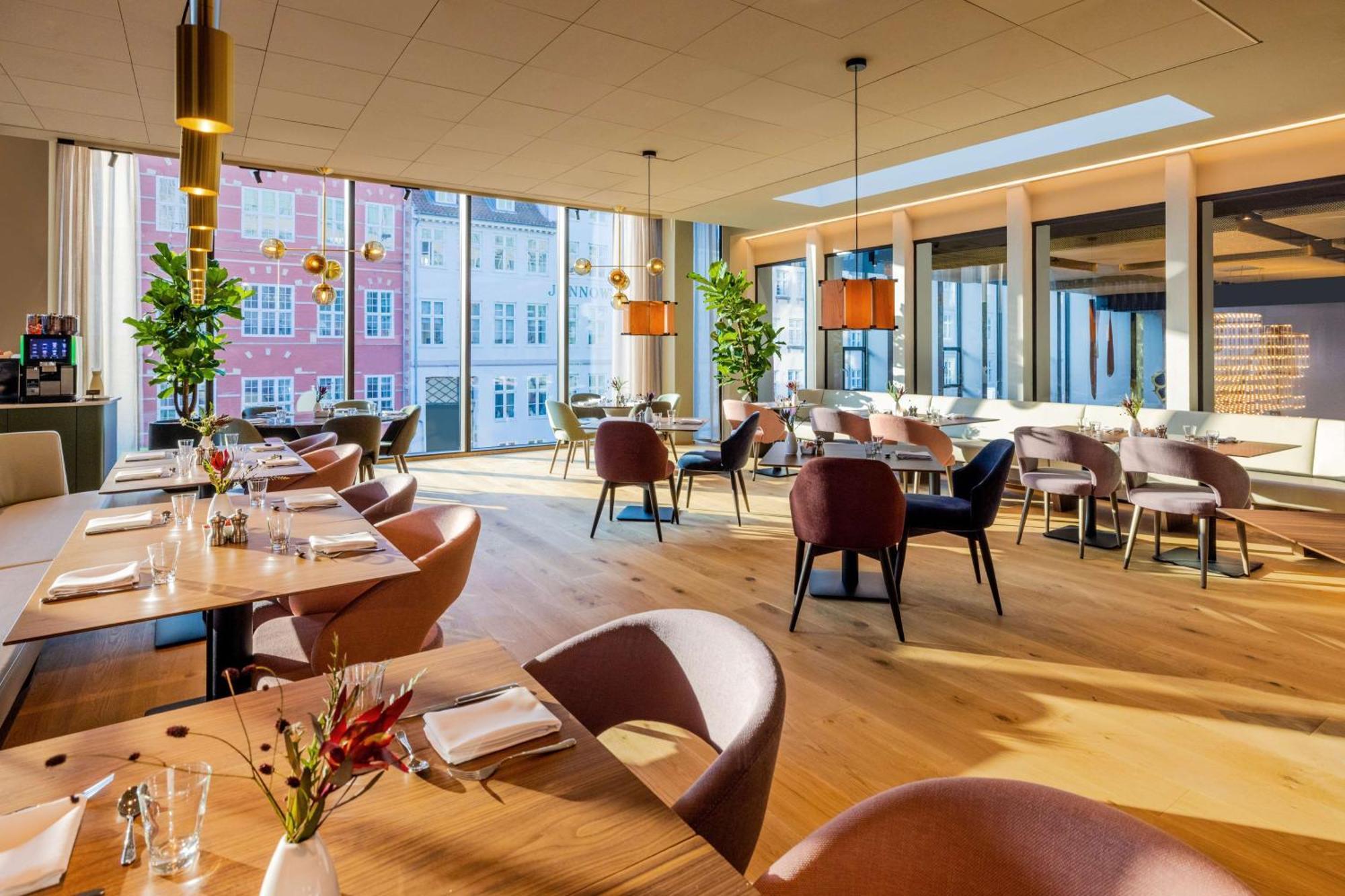 Nh Collection Copenhagen Hotel Ngoại thất bức ảnh The photo depicts a modern restaurant interior with ample natural light. The space features large windows that reveal buildings outside, enhancing the ambiance. Inside, there are several dining tables arranged with elegant table settings, including c